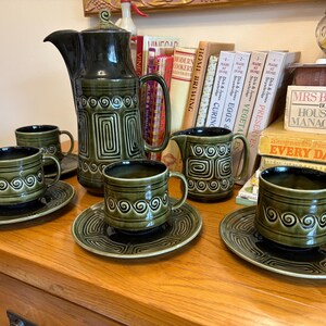 May include: A vintage green ceramic coffee set. The set includes a tall coffee pot, a creamer, and several cups with matching saucers. The design features geometric and swirl patterns. The cups and saucers are in a retro style.