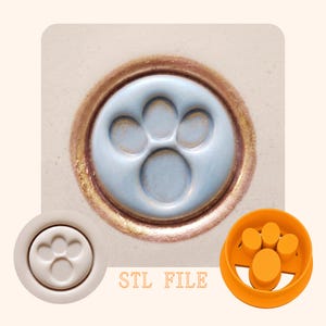 May include: A light blue paw print design within a gold-colored circular frame. The image also shows a white paw print and an orange paw print mold. The text "STL FILE" is displayed in the image.