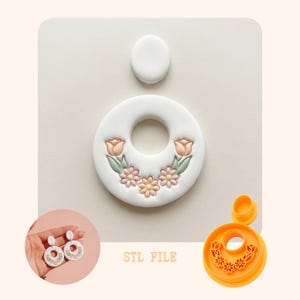 Op de afbeelding: Een wit donutvormig oorbelontwerp met een bloemmotief van roze tulpen en madeliefjes. De afbeelding bevat een bijpassende oorbel en een 3D-printmal. De tekst "STL FILE" is zichtbaar.
