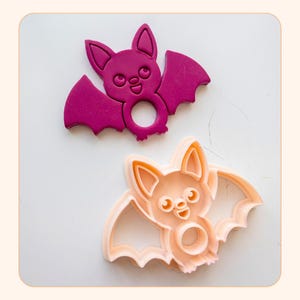 Pode incluir: Um cortador de biscoitos em forma de morcego na cor magenta escuro e um cortador de biscoitos em forma de morcego na cor pêssego claro. Ambos apresentam um design de morcego com orelhas pontudas, olhos grandes, nariz pequeno, boca aberta e asas estendidas com um recorte circular.