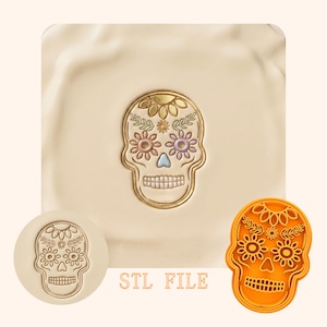May include: A gold-colored skull design with floral accents, set against a cream-colored background. The skull features detailed flower and leaf patterns. A round stamp and an orange plastic mold of the skull are also shown. The text "STL FILE" is visible.