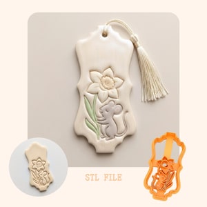 Puede incluir: Un marcapáginas crema con un diseño de narciso y ratón, con una borla. El diseño está en relieve en el marcapáginas. También se muestra un cortador de galletas naranja con el mismo diseño. El texto "STL FILE" es visible.