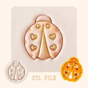 May include: A ladybird-shaped cookie cutter and stamp set featuring a pale pink ladybird with gold heart accents on its wings. The set includes a white circular stamp showing the ladybird outline and a bright orange plastic cutter. Text reads STL FILE.