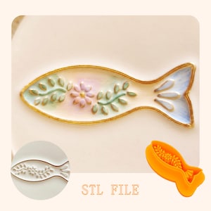 Könnte beinhalten: Ein fischförmiger Ausstecher aus leuchtend orangefarbenem Kunststoff, begleitet von einer weißen Keramikschale mit einem bemalten Fischmotiv, grünen Blättern, einer rosa Blume und Goldrand. Der Text STL FILE ist unten mittig in Orange gedruckt.