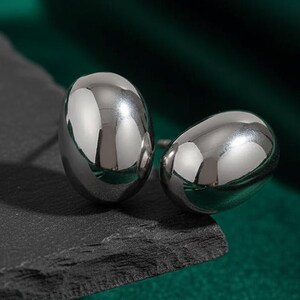 May include: A pair of polished, silver-colored stud earrings. The earrings have a smooth, oval shape and reflect the surrounding light. They are displayed on a dark gray, textured surface against a backdrop of deep green fabric.