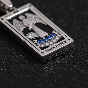 May include: A silver pendant featuring a tarot card design. The rectangular pendant has a black background with an angel illustration. The bottom of the pendant has the word "TEMPERANCE" and is adorned with blue and white gemstones.