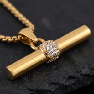 Peut inclure: Un collier doré avec un pendentif en forme de barre en T. Le pendentif est composé d'une barre cylindrique avec une section incrustée de petites pierres précieuses transparentes. La chaîne est de type maille carrée. Le collier est présenté sur une surface sombre et texturée.