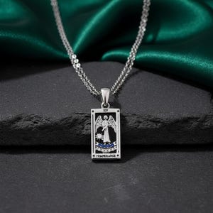 May include: A silver-toned necklace featuring a rectangular pendant with a tarot card design. The pendant depicts an angel figure with wings, holding a vessel. The card is labeled "XIV TEMPERANCE +" and has blue and white accents. The necklace hangs against a dark background.