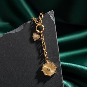 May include: A gold necklace featuring a heart-shaped charm and a circular pendant. The necklace has a chain with a toggle clasp and a long, dangling chain with a circular pendant. The pendant has a starburst design and is decorated with small, clear stones.
