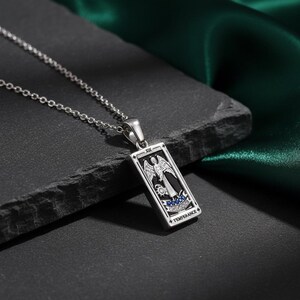 May include: A silver-toned pendant necklace featuring a rectangular charm with a detailed illustration of an angel. The charm has black and white accents, with blue embellishments and the word "TEMPERANCE". The necklace is displayed on a dark surface.