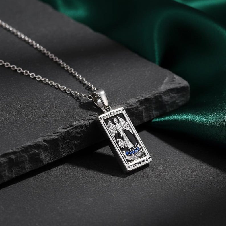 May include: A silver-toned pendant necklace featuring a rectangular charm with a detailed illustration of an angel. The charm has black and white accents, with blue embellishments and the word "TEMPERANCE". The necklace is displayed on a dark surface.