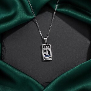 May include: A silver-toned necklace featuring a rectangular pendant with a tarot card design. The pendant depicts an angel with wings, blue accents, and the word "TEMPERANCE". The necklace hangs against a dark background with a green satin fabric.
