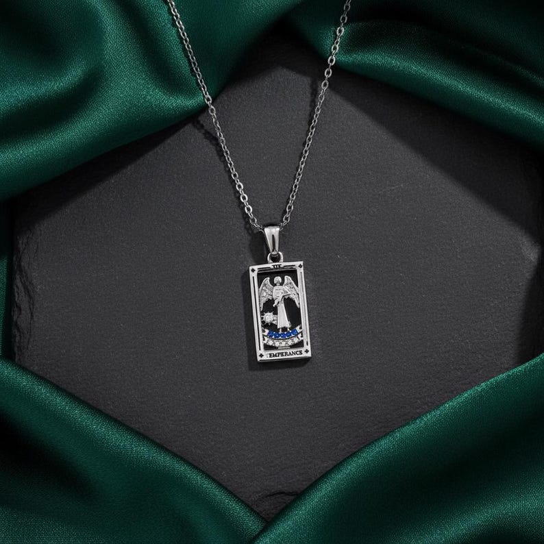 May include: A silver-toned necklace featuring a rectangular pendant with a tarot card design. The pendant depicts an angel with wings, blue accents, and the word "TEMPERANCE". The necklace hangs against a dark background with a green satin fabric.
