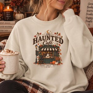 Può includere: Una felpa girocollo color crema con una stampa grafica di una vetrina di un caffè con il testo THE HAUNTED Cafe, COFFEE SPELLS BOO e EST. 1987. Il design include un fantasma, un gatto nero, zucche e foglie autunnali dai toni caldi.