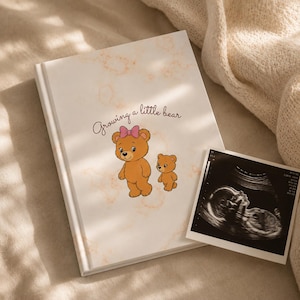 Può includere: Un libro con copertina rigida bianca con un motivo marmorizzato arancione chiaro, che riporta il testo "Growing a little bear" e un'illustrazione di due orsi bruni, uno dei quali con un fiocco rosa. Un'ecografia in bianco e nero è accanto al libro.