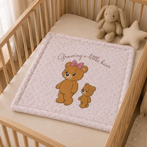 Può includere: Una coperta per neonati quadrata rosa chiaro con un bordo morbido e testurizzato. Il centro presenta un'illustrazione stampata di un grande orso marrone con un fiocco rosa e un orso più piccolo, accompagnata dal testo "Growing a little bear".