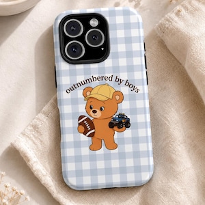 May include: A light blue and white gingham patterned phone case featuring a brown teddy bear wearing a yellow cap. The bear holds a brown football and a blue toy truck. The text "outnumbered by boys" is printed in brown above the bear.