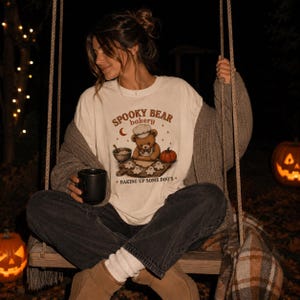 Può includere: Una t-shirt color bianco sporco con la grafica di un orsacchiotto marrone con cappello da chef che prepara biscotti a forma di fantasma. Il testo recita SPOOKY BEAR bakery e BAKING UP SOME BOO'S, con una luna crescente, stelle e una zucca.