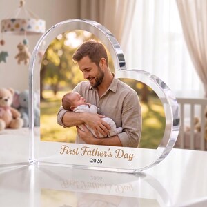 May include: A heart-shaped clear acrylic photo frame with a picture of a man holding a baby. The text "First Father's Day 2026" is printed in gold script at the bottom of the frame. The frame is on a white surface.
