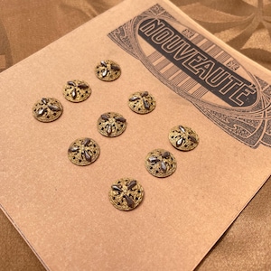 Puede incluir: Diez botones dorados antiguos con un diseño floral están dispuestos sobre un papel marrón claro con la palabra "NOUVEAUTÉ" impresa en negro. Los botones tienen un centro oscuro y detalles intrincados.