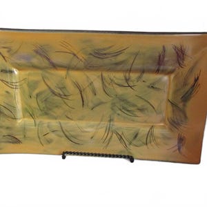 May include: A rectangular ceramic serving tray featuring a warm mustard yellow and tan base colour. The surface is decorated with dark brown and black brushstroke patterns that create a series of curved, linear marks across the entire tray.