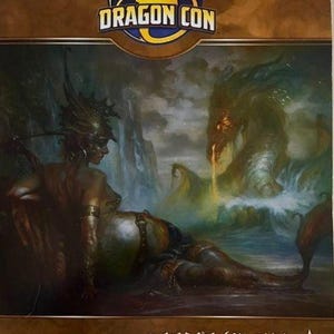 Può includere: Un libretto del programma Dragon Con 2014 con un'illustrazione fantasy di una figura dalla pelle scura in armatura ornata sdraiata vicino a un grande drago marino dagli occhi luminosi che emerge dall'acqua. Il testo recita Dragon Con e Program Book.