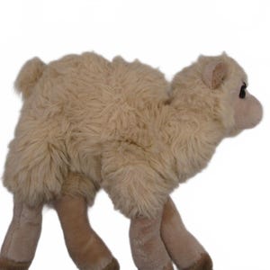 May include: A plush camel toy featuring a shaggy, light beige coat with a soft, textured appearance. The stuffed animal has a rounded hump, four legs with darker brown hooves, and a small head with dark eyes and rounded ears facing to the right.
