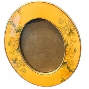 May include: An oval picture frame with a bright yellow border featuring a floral pattern of yellow and green flowers. The frame has a metallic gold-toned inner and outer edge, surrounding a plain, dark brown oval-shaped center insert.