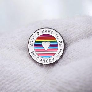 Peut inclure: &Eacute;pingle ronde en &eacute;mail avec une bordure blanche et un texte noir qui dit "YOU'RE SAFE TO BE YOU AROUND ME". L'&eacute;pingle pr&eacute;sente un motif ray&eacute; aux couleurs de l'arc-en-ciel avec un c&oelig;ur blanc au centre.