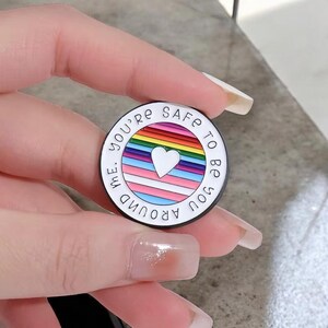 Peut inclure: Un support de t&eacute;l&eacute;phone rond, blanc et noir, avec le texte "You're safe to be you around me." Le centre pr&eacute;sente un arc-en-ciel, un c&oelig;ur blanc et le drapeau de la fiert&eacute; transgenre.