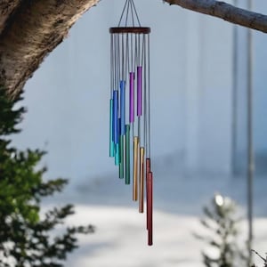 May include: A rainbow-colored wind chime hangs from a tree branch. The chime features a dark wooden top and a series of colorful metal tubes in shades of blue, green, purple, gold, and red. The tubes are suspended by thin black cords.