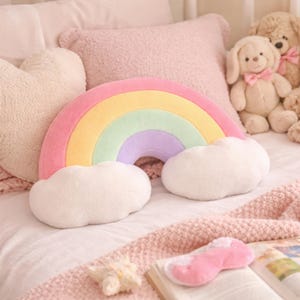 May include: A colourful rainbow-shaped cushion with pink, yellow, green, and purple arches, and white cloud details. The cushion is on a bed with other pillows and soft toys. The overall colour scheme is soft pastels.