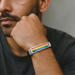 May include: A silver bracelet with a rainbow-colored rectangular design. The bracelet has a black band and is worn on the wrist. The rainbow design features stripes of red, orange, yellow, green, blue, and purple.