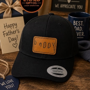 May include: A black baseball cap with a mesh back and a rectangular tan leather patch on the front. The patch is laser-engraved with the word DADDY in a handwritten font. A silver and black sticker on the brim reads THE CLASSICS SNAPBACK.