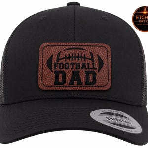 Puede incluir: Una gorra de béisbol negra estilo camionero con un parche rectangular de piel sintética marrón con textura de balón de fútbol. El parche muestra el texto FOOTBALL DAD en letras negras. Un logotipo circular indica ETCHED GIFT CO.
