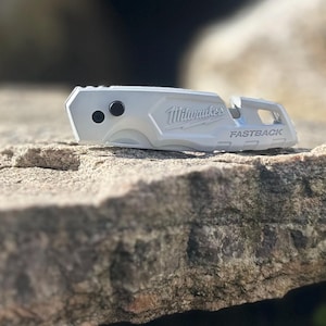 May include: A silver Milwaukee Fastback utility knife featuring a metallic finish, two black circular buttons on the side, and the brand name Milwaukee and the word FASTBACK embossed on the handle, shown in a closed, compact position.