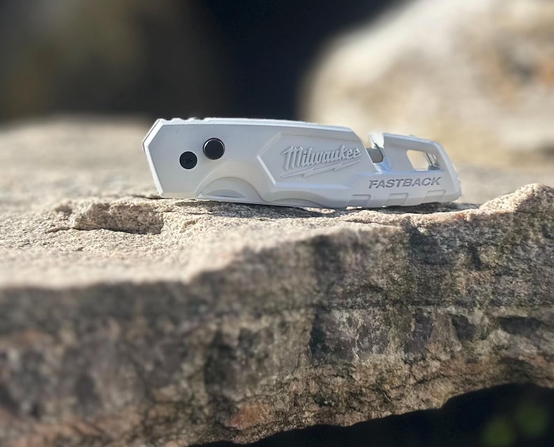 May include: A silver Milwaukee Fastback utility knife featuring a metallic finish, two black circular buttons on the side, and the brand name Milwaukee and the word FASTBACK embossed on the handle, shown in a closed, compact position.