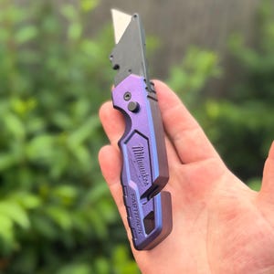 May include: A purple and black Milwaukee Fastback folding utility knife with a metallic finish. The tool features a sharp, exposed metal blade, a black pivot screw, and the brand name Milwaukee and the word FASTBACK embossed on the handle.
