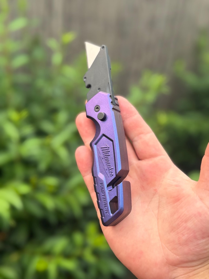 May include: A purple and black Milwaukee Fastback folding utility knife with a metallic finish. The tool features a sharp, exposed metal blade, a black pivot screw, and the brand name Milwaukee and the word FASTBACK embossed on the handle.