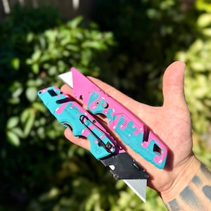 May include: Two Milwaukee Fastback utility knives featuring a custom pink and teal donut-themed paint pattern. The knives have metal blades, black belt clips, and the Milwaukee logo printed on the handles with model number 48-22-1502 visible.