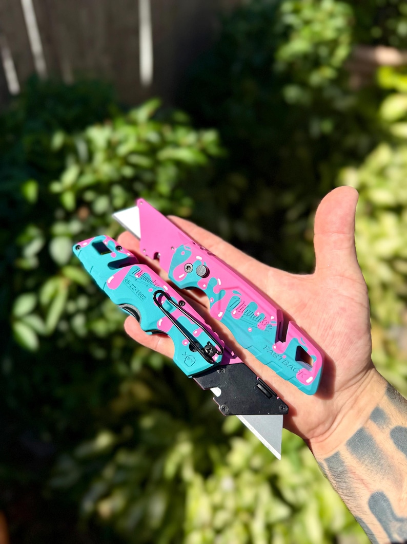 May include: Two Milwaukee Fastback utility knives featuring a custom pink and teal donut-themed paint pattern. The knives have metal blades, black belt clips, and the Milwaukee logo printed on the handles with model number 48-22-1502 visible.