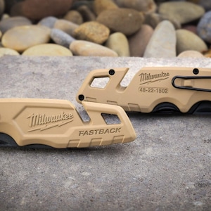May include: Two tan Milwaukee Fastback folding utility knives. The left knife features the Milwaukee logo and FASTBACK text. The right knife displays the Milwaukee logo, model number 48-22-1502, a black metal pocket clip, and a small blade icon.