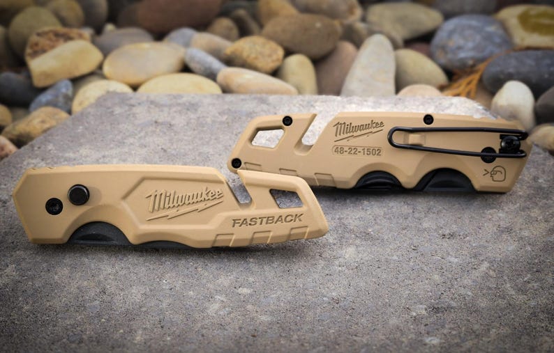 May include: Two tan Milwaukee Fastback folding utility knives. The left knife features the Milwaukee logo and FASTBACK text. The right knife displays the Milwaukee logo, model number 48-22-1502, a black metal pocket clip, and a small blade icon.