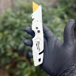 May include: A white Milwaukee Fastback utility knife with a gold-coloured blade extended. The handle features the Milwaukee logo and the word Fastback printed in black text. The tool has a folding design with a metal frame and a belt clip slot.