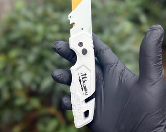 以下が含まれることがあります： ゴールドカラーのブレードが伸びた白いMilwaukee Fastbackユーティリティナイフ。ハンドルにはMilwaukeeのロゴとFastbackという文字が黒でプリントされています。金属フレームとベルトクリップを備えた折りたたみ式のデザインです。