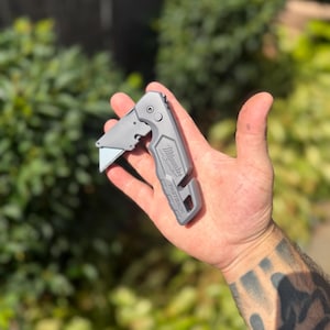 May include: A grey Milwaukee Fastback folding utility knife with a metal body, a partially extended silver blade, a locking mechanism, and a cutout at the base. The brand name Milwaukee and the model name Fastback are embossed on the handle.