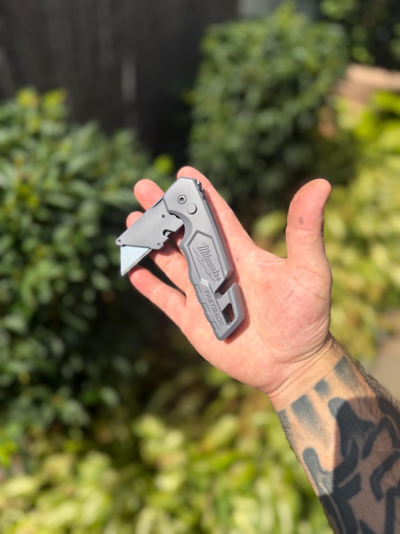 May include: A grey Milwaukee Fastback folding utility knife with a metal body, a partially extended silver blade, a locking mechanism, and a cutout at the base. The brand name Milwaukee and the model name Fastback are embossed on the handle.