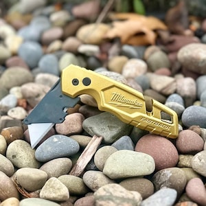 May include: A gold-coloured Milwaukee Fastback utility knife with a partially extended silver blade. The handle features the Milwaukee logo and the word Fastback embossed on the side, with a black pivot point and a metallic finish.