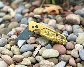 以下が含まれることがあります： ゴールドカラーのMilwaukee Fastbackユーティリティナイフ。シルバーのブレードが一部露出しています。ハンドル側面にはMilwaukeeのロゴとFastbackの文字が刻印されており、黒いピボットポイントとメタリックな仕上げが施されています。