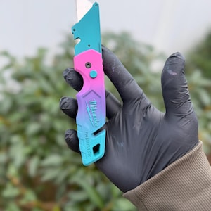 May include: A Milwaukee Fastback utility knife featuring a vibrant teal and pink gradient finish. The folding tool has a metallic blade extended at the top, a central release button, and the brand name printed on the handle.